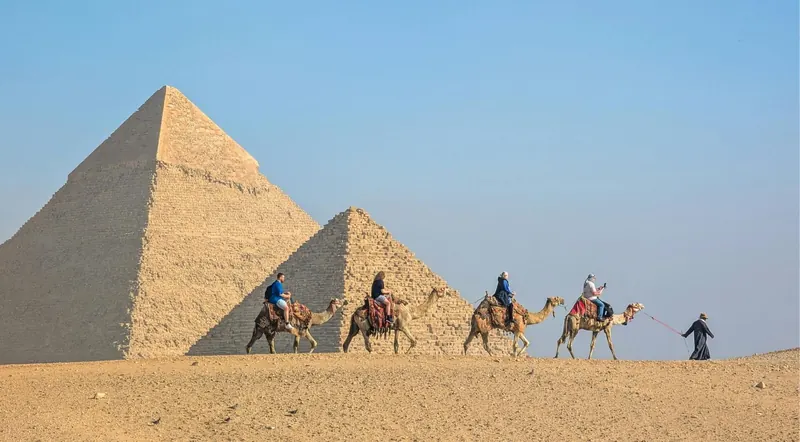 Tourists riding camels in a caravan past the pyramids of Giza